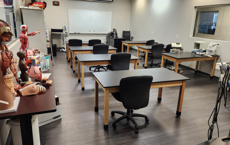 Medical classroom with models and charts, black chairs and tables, whiteboard, and various equipment.