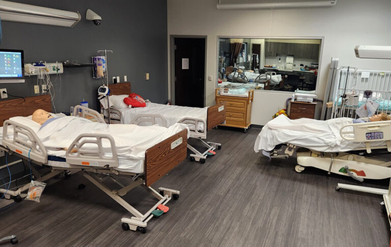 Hospital room with three beds, two containing mannequins. Medical equipment and supplies are visible around the room.