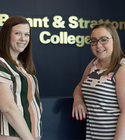 Two women smiling and standing in front of a sign that reads "Bryant & Stratton College." One wears a striped shirt, while the other has a floral pattern. They appear friendly and welcoming.