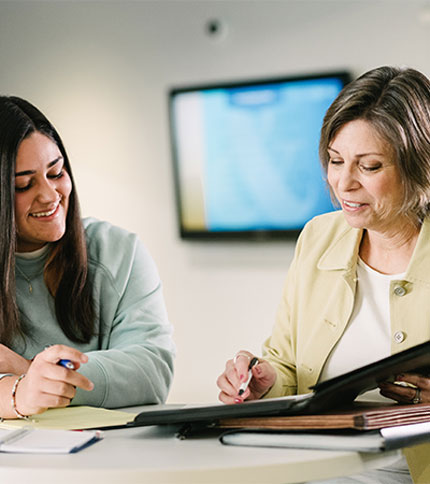 Two women sit at a table, smiling while reviewing documents. One woman is writing with a pen, and the other is holding a folder. There's a blurred screen in the background.