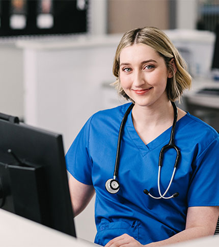 A healthcare professional in blue scrubs and a stethoscope smiles while sitting at a computer in a clinical setting.