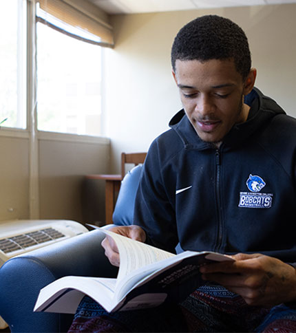 A person in a black "Bobcats" hoodie reads a book about online medical billing and coding programs while sitting in a cozy chair by a softly lit window. The room exudes a relaxed atmosphere, perfect for studying or unwinding with new knowledge.