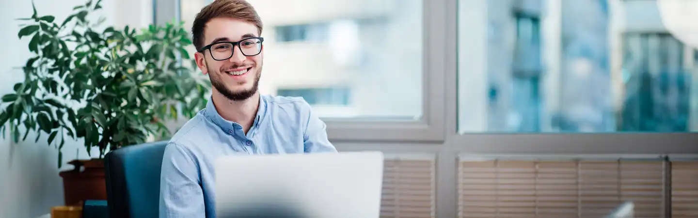 A smiling man with glasses is sitting at a desk in front of a laptop, with a plant and large windows in the background.