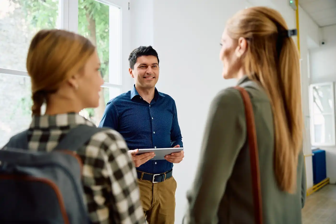 A man holding a tablet smiles at two women, one with a backpack, inside a well-lit room with large windows.