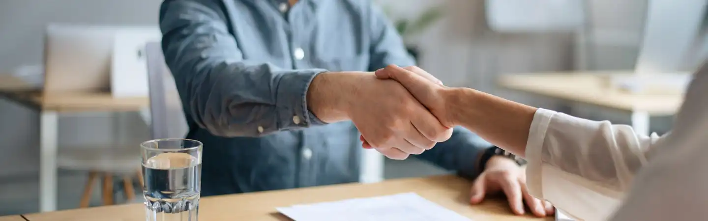 Two people shaking hands across a desk with a document and a glass of water in the foreground.