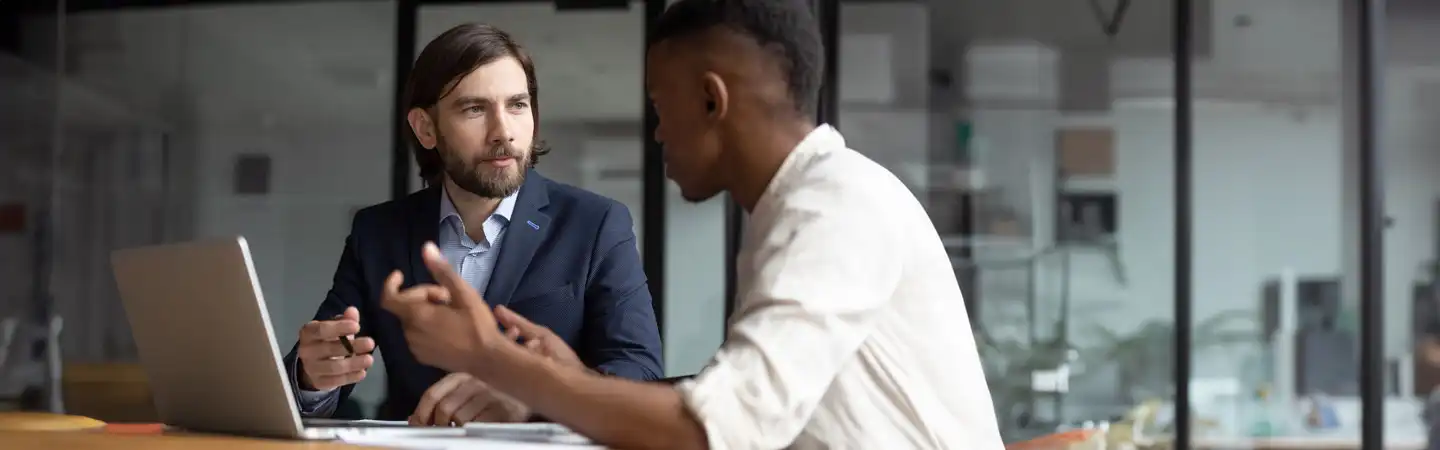 Two men in an office setting, discussing something at a desk with an open laptop.