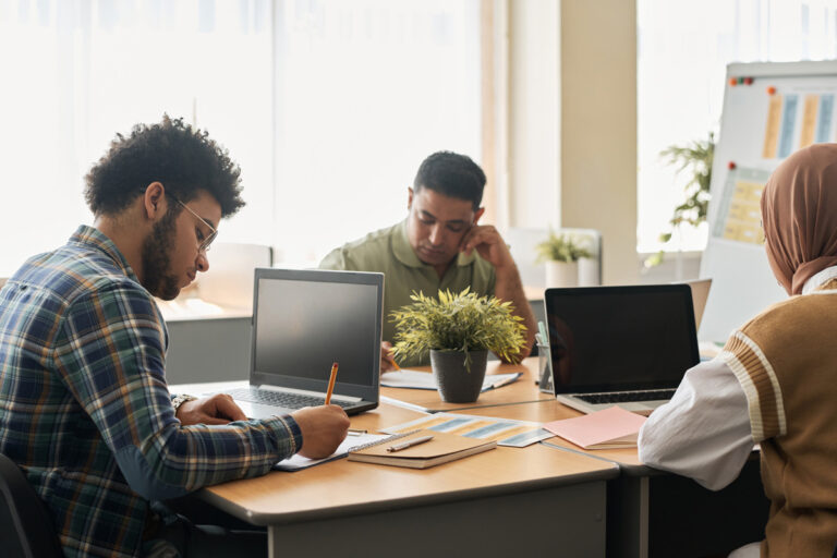 Three people working at laptops around a table with plants, taking notes. One person wears a hijab.