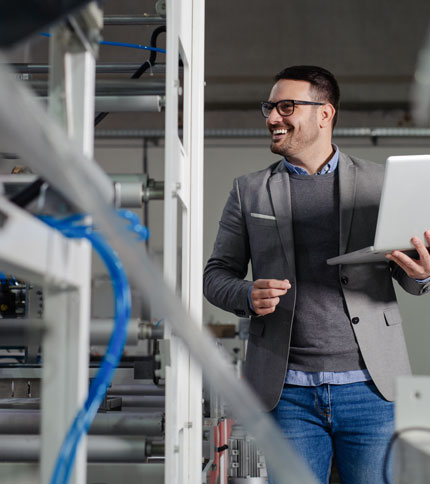 A smiling man wearing glasses and a gray blazer holds a laptop while standing in an industrial setting with machinery around.