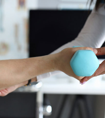 A person in a white shirt hands a small blue hexagonal dumbbell to another person. They are in a bright indoor setting, possibly a gym or physical therapy room.