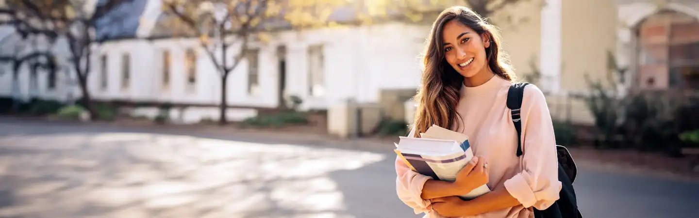 female student with a backpack holding books smiling at camera on a college campus