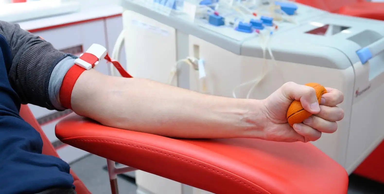 Person donating blood, lying on a red chair, squeezing a stress ball, with medical equipment in the background.