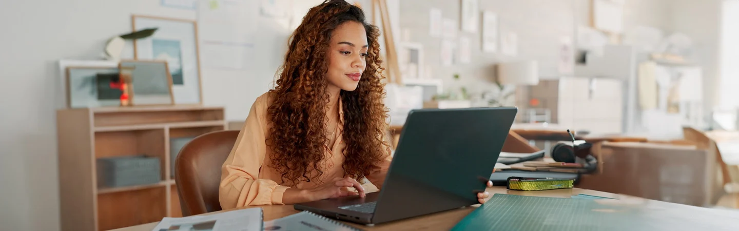 Woman with curly hair working on a laptop at a desk in a well-lit office space.