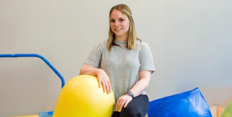 A person smiling, sitting next to a large yellow exercise ball, with blue and green soft play equipment in the background.