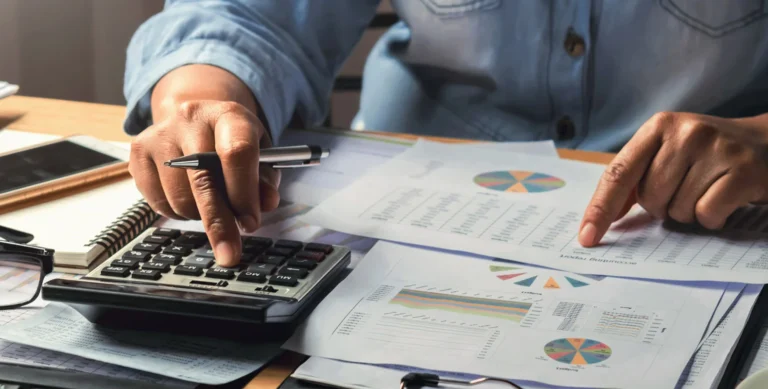 Person using a calculator and reviewing financial charts and graphs on a desk, wearing a casual shirt.