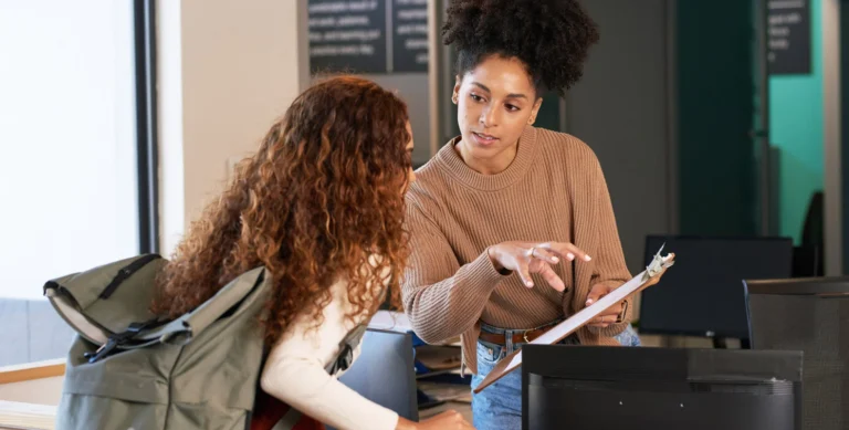 Two women engaged in a conversation at an office desk, one holding a clipboard while explaining something to the other.