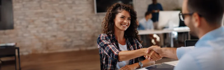 A smiling woman with curly hair shakes hands with a man in an office setting.