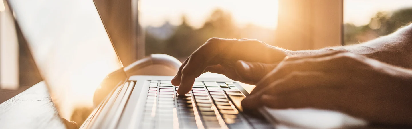 Close-up of hands typing on a laptop keyboard with sunlight streaming through a window in the background.