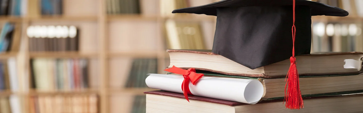 Graduation cap and diploma with a red ribbon on top of stacked books in front of a blurred bookshelf background.