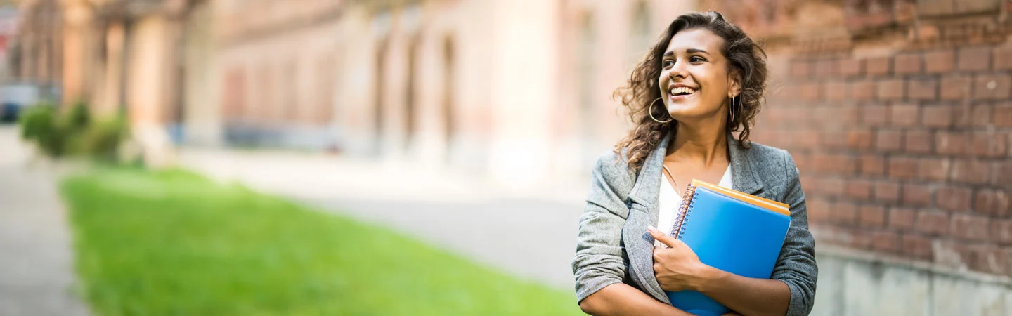 A woman smiles while holding notebooks, standing outdoors near a brick building and green lawn.