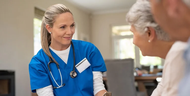 A smiling healthcare worker in blue scrubs talks to a senior couple inside a home setting.