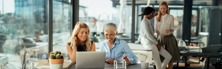 Two women working together on a laptop at a desk, with two more people conversing in the background in an office setting.