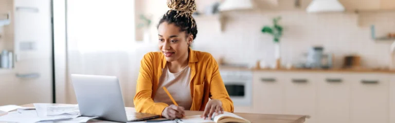 Woman in a yellow jacket working on a laptop and taking notes at a kitchen table.