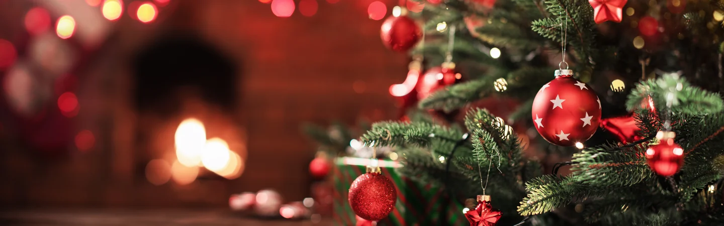 Close-up of a decorated Christmas tree with red baubles and lights, a cozy fireplace glowing in the background.