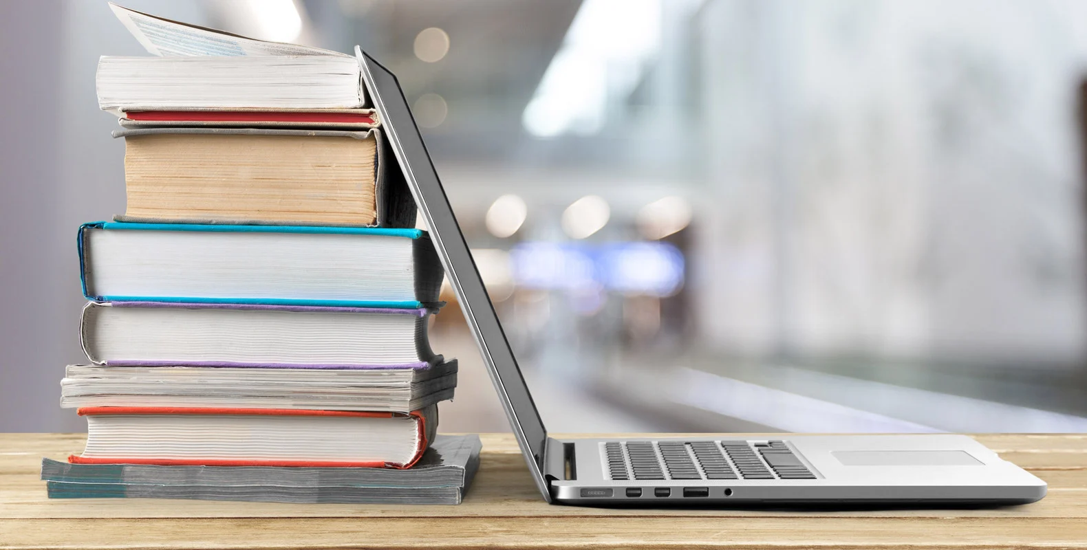 A stack of books next to an open laptop on a desk in a blurred, modern indoor setting.