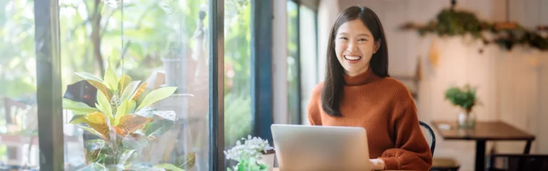 A smiling woman in a brown sweater sits by a large window, working on a laptop in a cozy, plant-filled cafe.