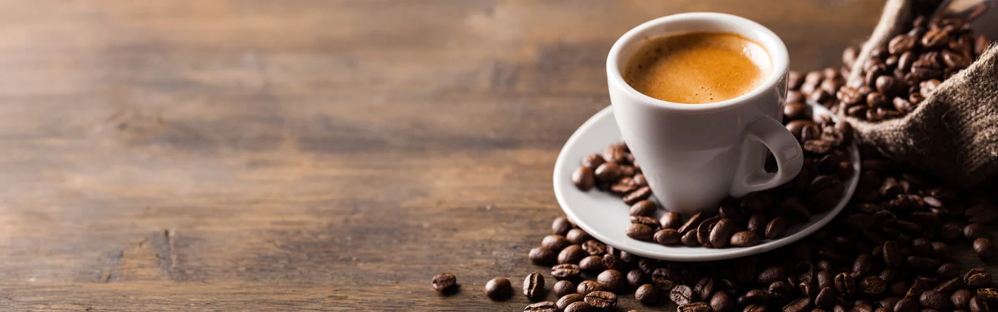 A cup of espresso on a saucer surrounded by coffee beans, with some spilling from a burlap sack on a wooden surface.