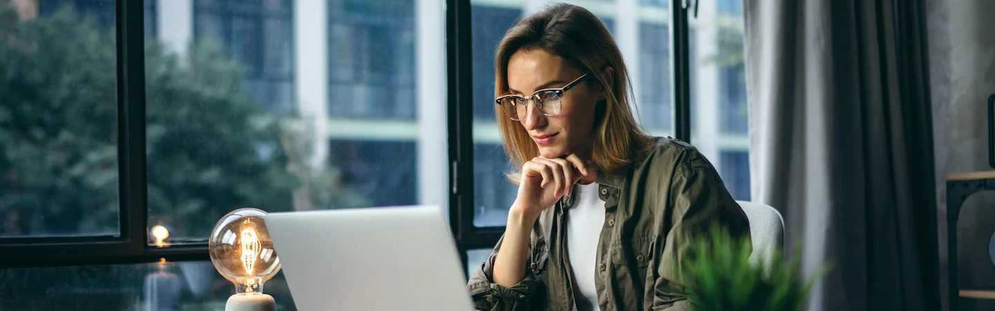 Woman with glasses working on a laptop at a desk near a window with a light bulb lamp and a potted plant.