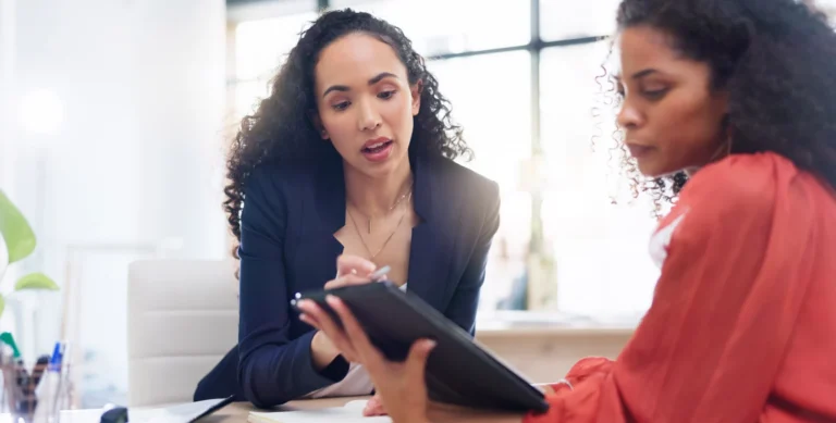 Two women with curly hair in a professional setting, one showing the other a document on a clipboard.