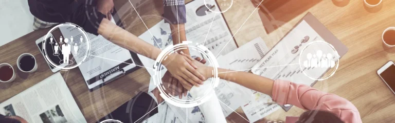 Top view of hands stacked in the center over a table with documents and infographics, symbolizing teamwork and collaboration.