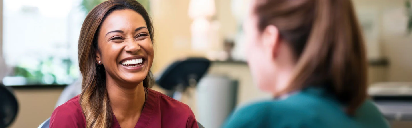 Two women, one in maroon scrubs and the other in green, share a laugh in a well-lit room.