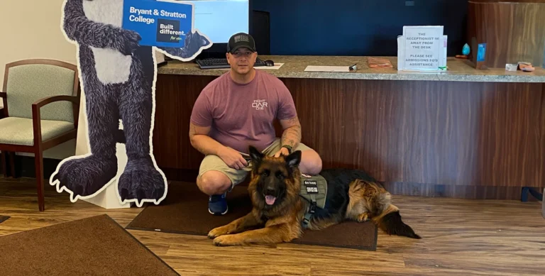 A man kneels next to a German Shepherd dog wearing a vest inside an office lobby with signs and chairs in the background.