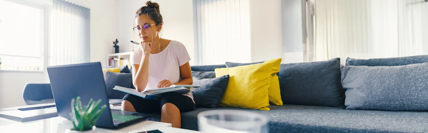 A woman with glasses working on a laptop at home, sitting on a gray sofa with yellow cushions, holding a notebook.