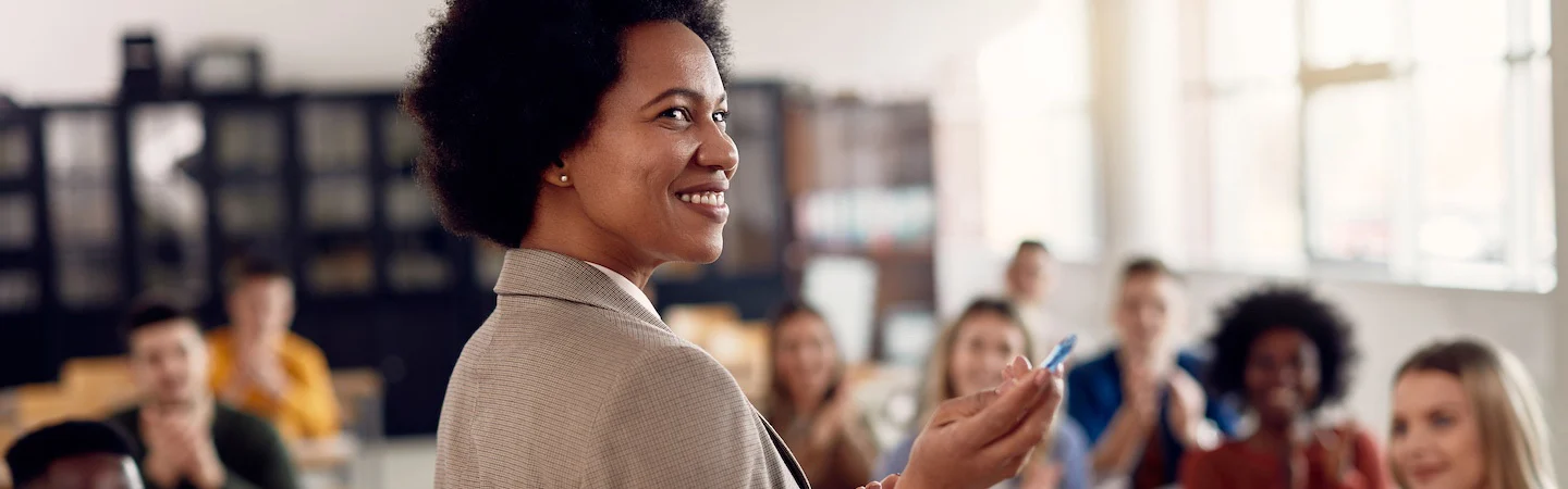 Smiling person in a blazer holds a marker while standing in front of a clapping audience in a bright, classroom-like setting.