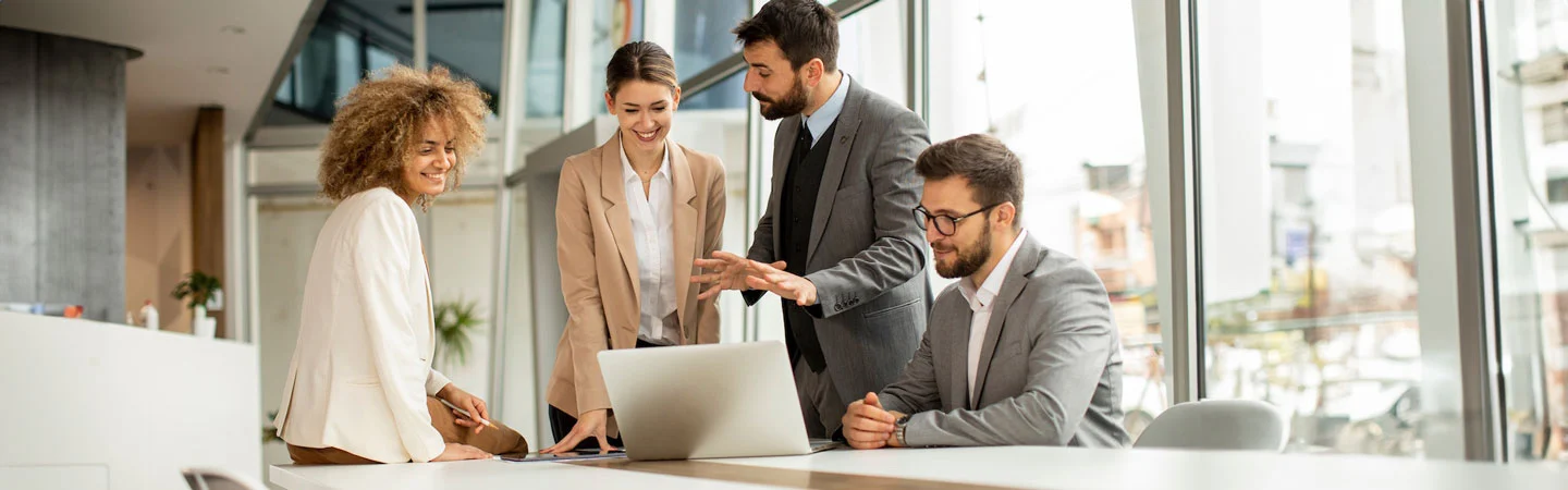 Four professionals in business attire enthusiastically discussing a project around a laptop in a modern office.