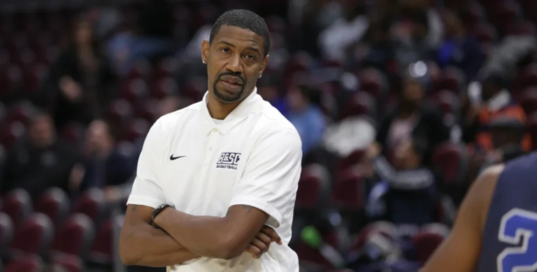 A man in a white polo shirt stands with arms crossed in a basketball stadium, looking towards the court.