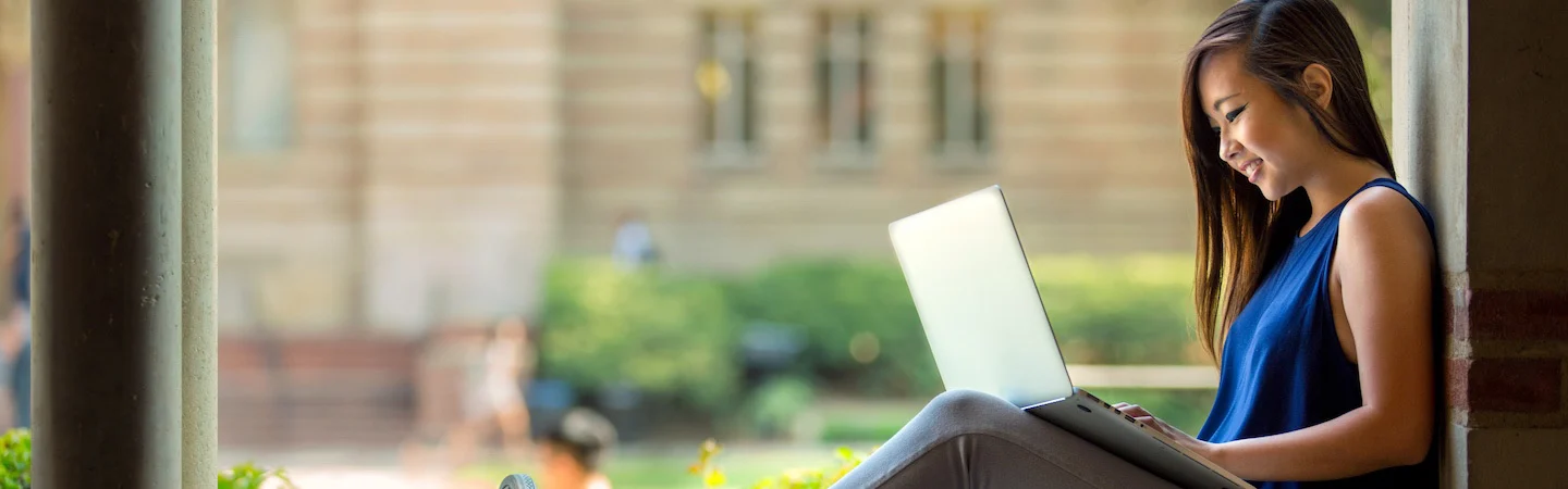 A young woman in casual clothing sits outdoors, using a laptop and smiling. A campus building is blurred in the background.