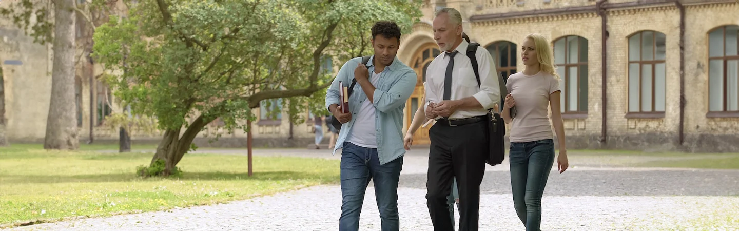 Three people walking on a university campus, with one person in a suit holding a briefcase, and the other two casually dressed.