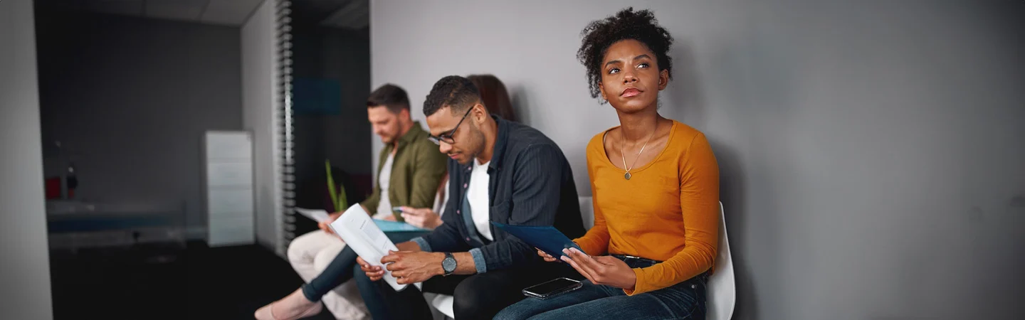 Four people sit on chairs in a waiting room, holding documents and devices, with one person looking at the camera.