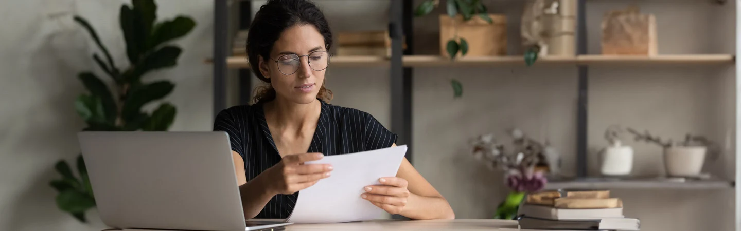 Person wearing glasses, sitting at a desk with a laptop, reading a document in a well-lit room with shelves in the background.