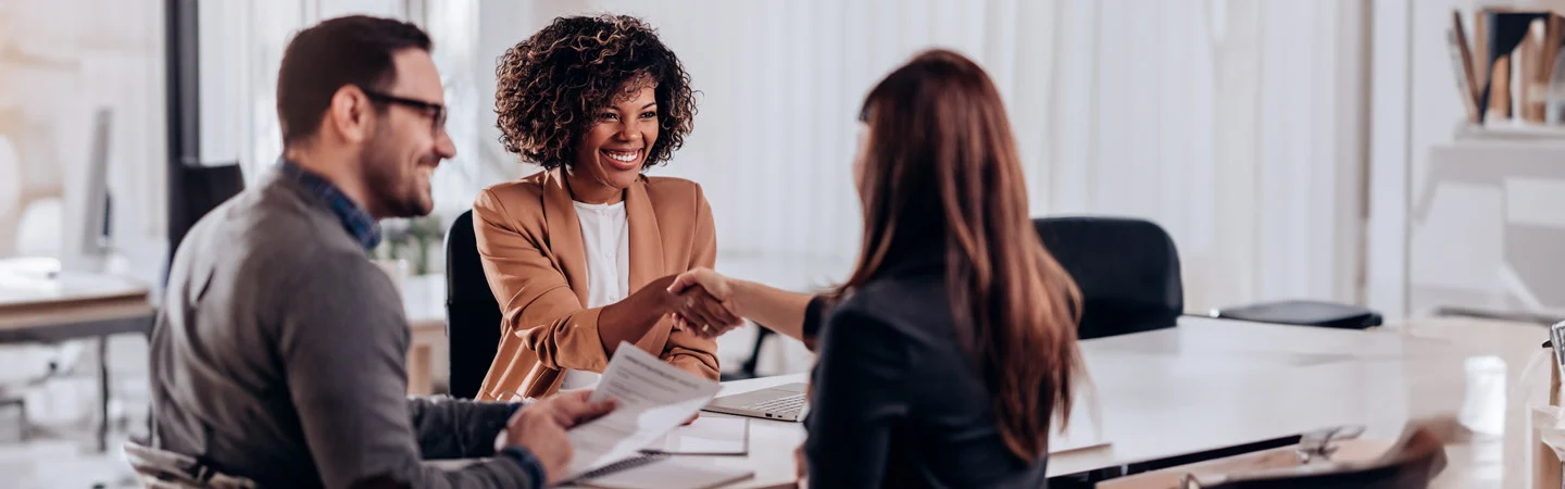 Three people in an office, two women shaking hands and a man holding papers, suggesting a business meeting or interview.