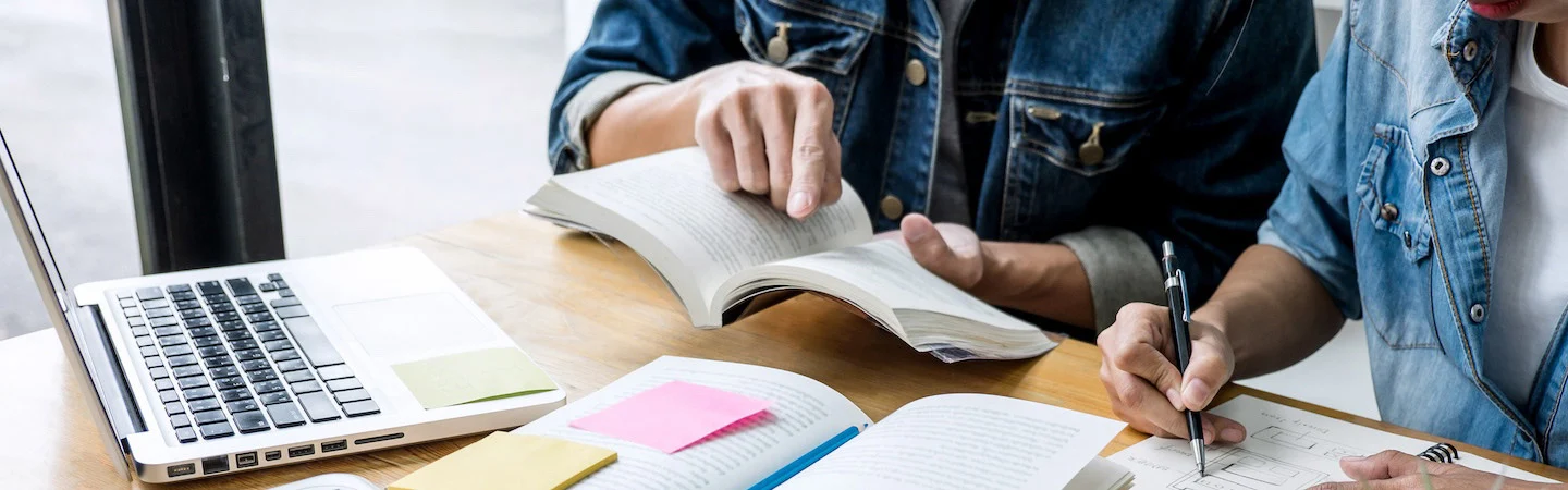 Two people studying together with open books, a laptop, and notes on a wooden table.