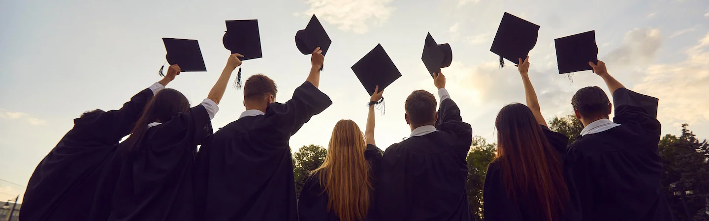 Graduates in caps and gowns hold their mortarboards in the air, celebrating outdoors against a cloudy sky.