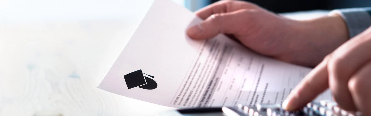 Close-up of a person holding a document with a graduation cap icon, while typing on a calculator.