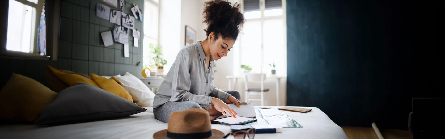 Woman sitting on bed, working with papers and laptop in a well-lit, cozy bedroom.