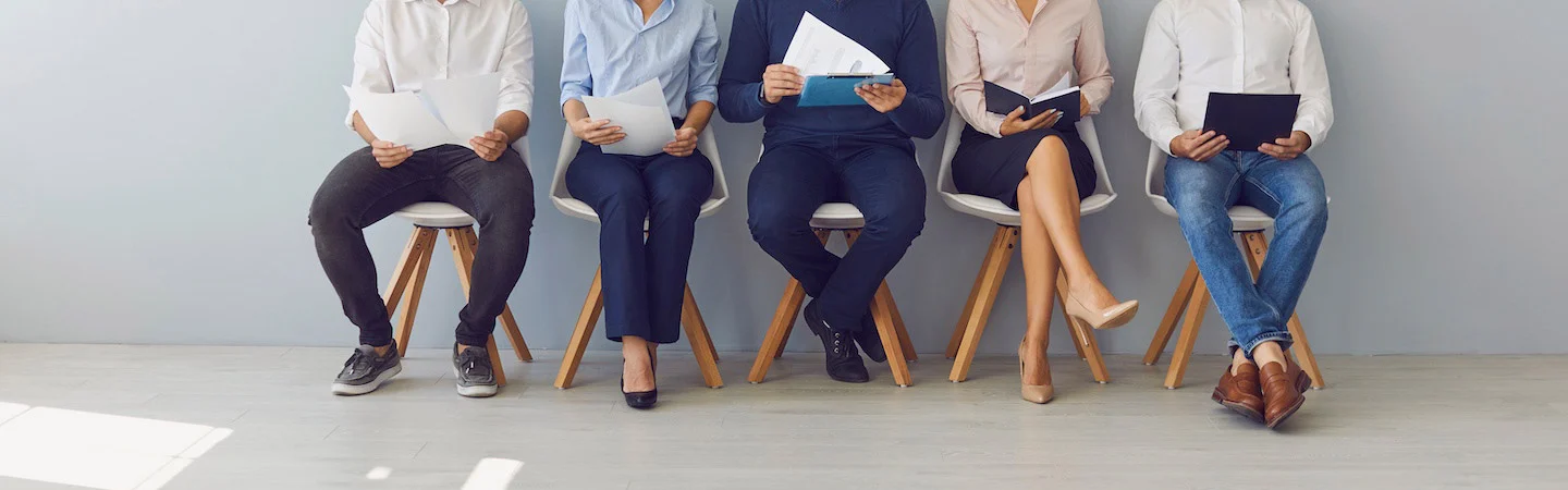 Five people sitting in chairs, holding papers, likely waiting for a job interview in a bright room with a gray wall.