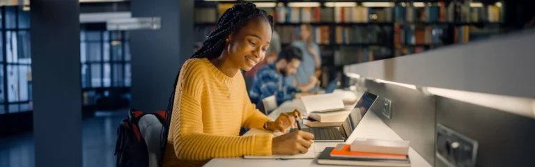 A person in a yellow sweater studies in a library, writing in a notebook and using a laptop.